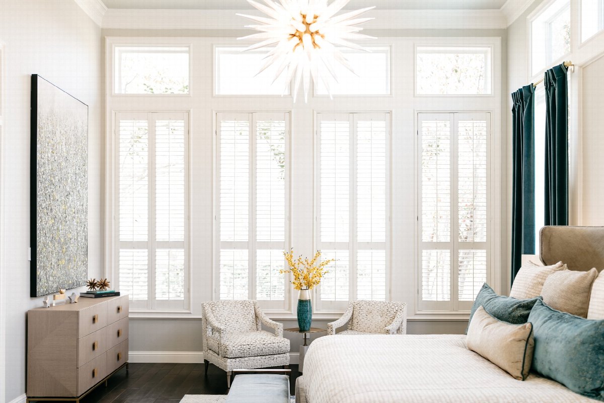 Luxury bedroom with statement chandelier, upholstered bed, accent chairs, and large windows in a bright, elegant interior design.