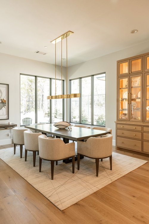 Luxury dining room with modern chandelier, upholstered chairs, wood table, and large windows in a bright, contemporary interior.
